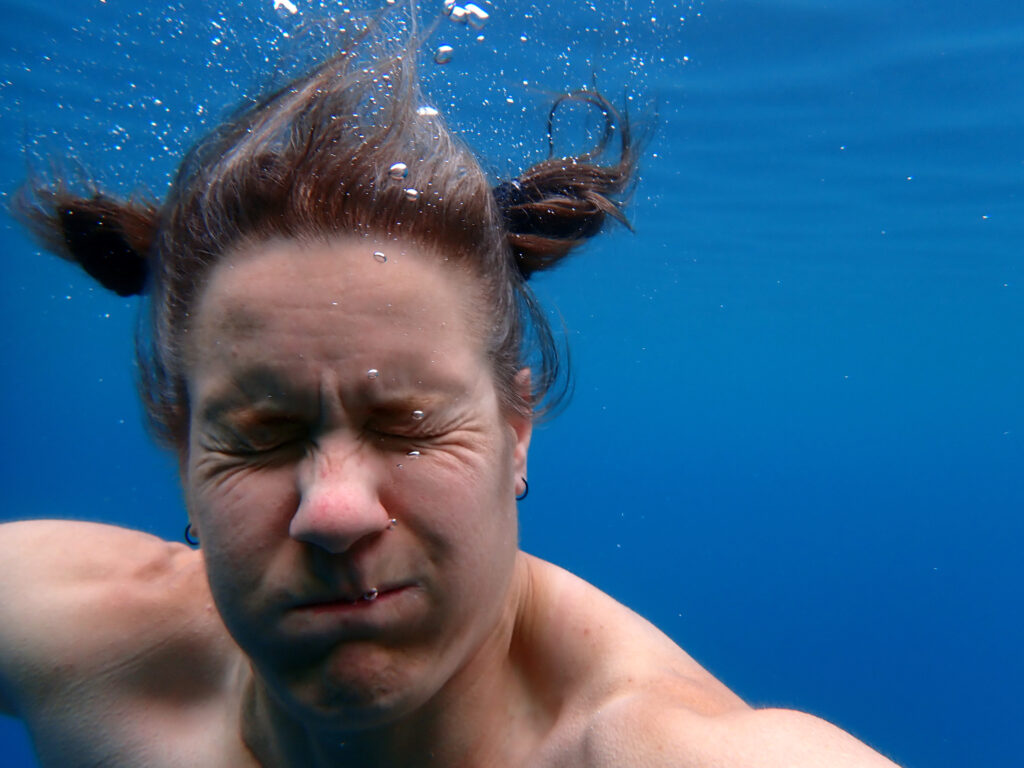 A woman holding her breath under water. Her eyes are shut tight and there are some small bubbles